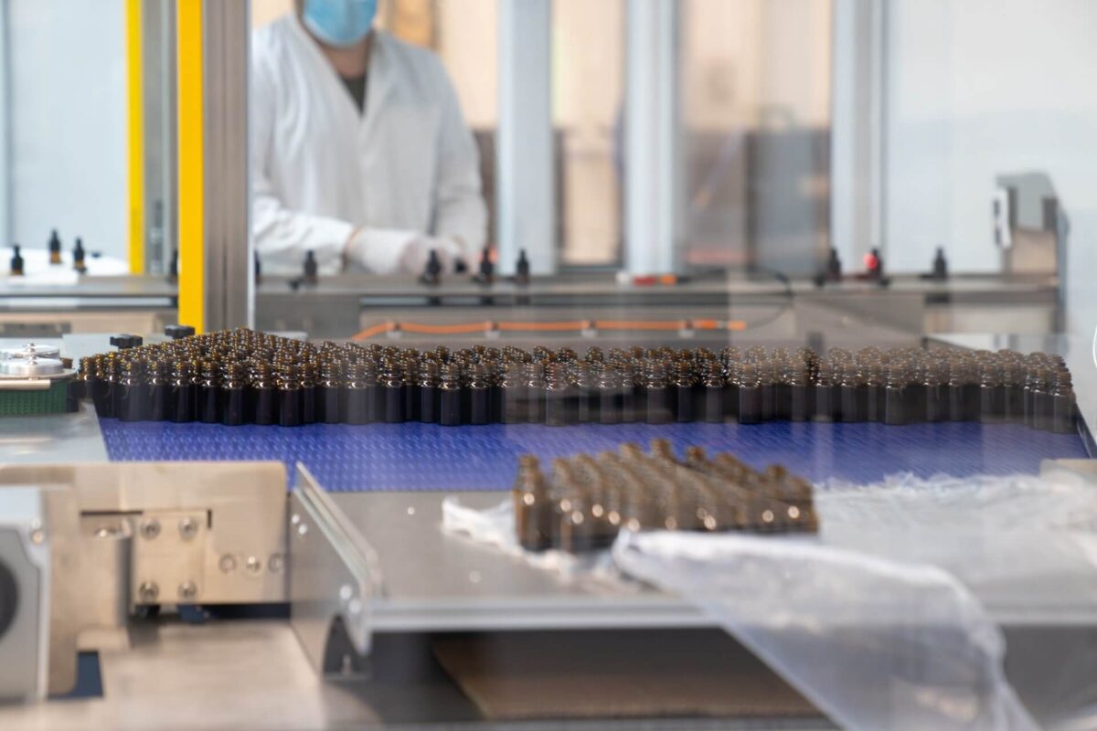 Ein Fabrikarbeiter in weißem Laborkittel und Gesichtsmaske arbeitet in einer Reinraumumgebung. Im Vordergrund sind mehrere kleine, dunkel gefärbte CBD-Flaschen auf einem Förderband aufgereiht, wahrscheinlich Teil eines Herstellungs- oder Verpackungsprozesses.